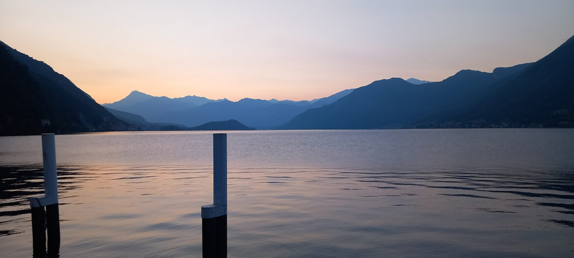 Morning lake view from Italian villa terrace at dawn: calm waters reflect Alps and wooden posts under soft pink sky, evoking unhurried rhythms of extended stays.